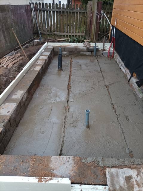 Świeżo wylana betonowa podłoga z wystającymi rurami instalacyjnymi, obramowana ceglanym fundamentem i drewnianym ogrodzeniem w tle.