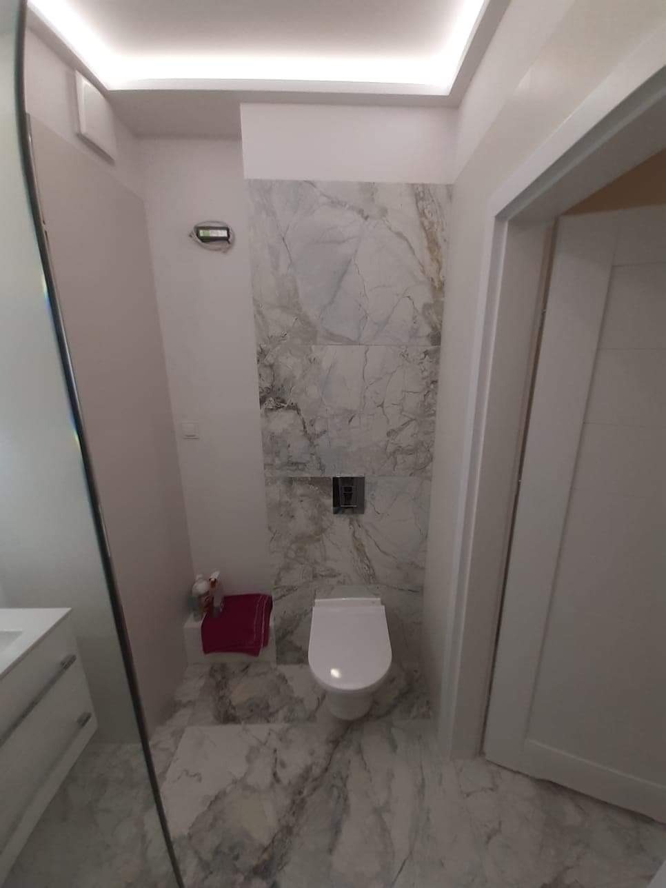 Wąska łazienka wykończona płytkami imitującymi marmur, z podwieszaną toaletą i oświetleniem LED w suficie podwieszanym.