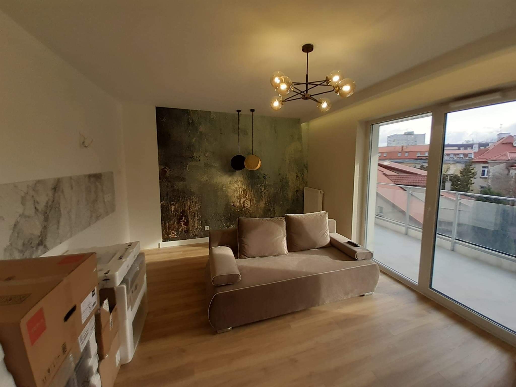 Nowoczesny salon z beżową sofą, abstrakcyjną tapetą w odcieniach zieleni i brązu, oraz designerską lampą sufitową z kulistymi kloszami. Duże okno balkonowe z widokiem na miejski krajobraz. Podłoga...