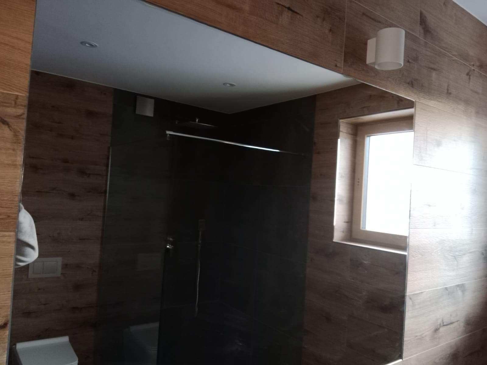 Nowoczesna łazienka z kabiną prysznicową typu walk-in, wykończona płytkami imitującymi drewno i ciemnymi płytkami w kabinie, oświetlona kinkietem i światłem dziennym z okna.