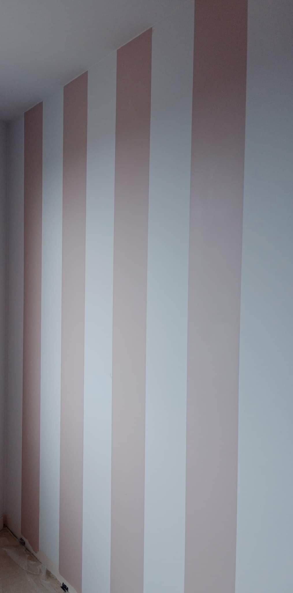 Pomalowana ściana w pionowe pasy w odcieniach pudrowego różu i bieli, widok z dołu ukazujący wzór na ścianie, świeżo po malowaniu.