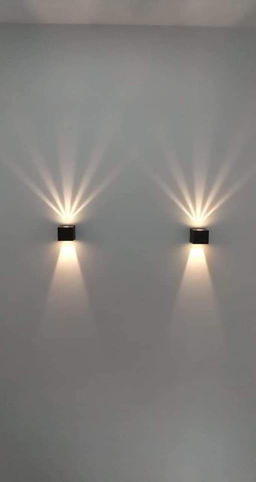 Dwa czarne, kwadratowe kinkiety ścienne emitujące promienie światła w górę i w dół na gładkiej, szarej ścianie.