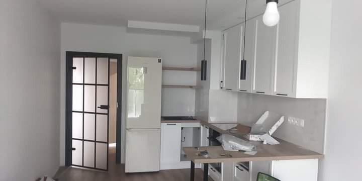 Wnętrze kuchni w trakcie montażu: białe szafki wiszące, lodówka, czarna stalowa rama drzwi z szybami, minimalistyczne lampy wiszące, blat roboczy z elementami opakowań.