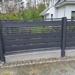 Nowoczesne, poziome ogrodzenie panelowe w kolorze grafitowym, z widocznym fragmentem domu w tle i elementami dekoracyjnymi z kory drzewnej u podstawy.