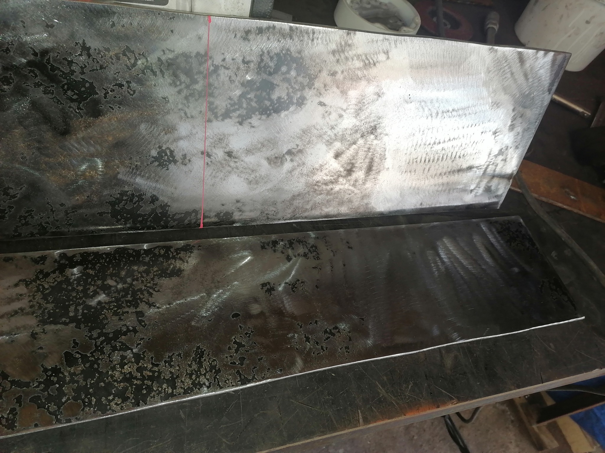 Dwie płyty metalowe o surowej powierzchni, z widocznymi śladami obróbki i zarysowaniami, leżące na warsztatowym stole, z czerwoną linią naniesioną na górnej płycie.