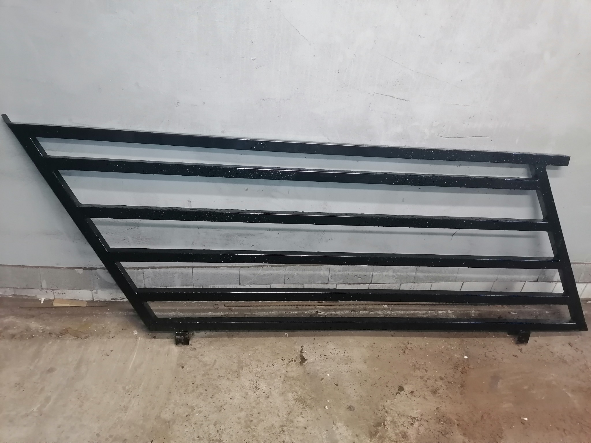 Czarna, metalowa konstrukcja bramy lub furtki, leżąca na podłodze w trakcie spawania lub malowania, z widocznymi elementami spawów i niedoskonałościami farby.