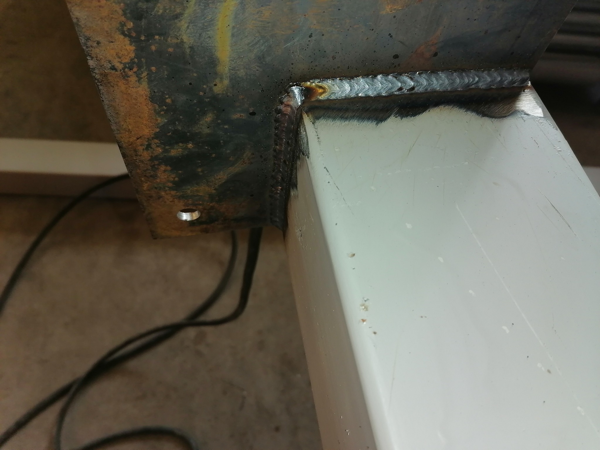 Spoina spawalnicza łącząca rdzewiejącą, czarną płytę z białą, kwadratową rurą; widoczne ślady użytkowania i niedokładne wykończenie.