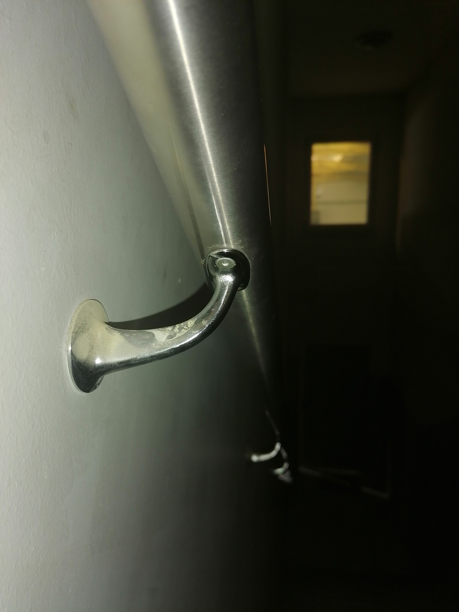 Fragment ściany z zamontowaną, metalową poręczą w korytarzu, widoczny uchwyt mocujący poręcz do ściany i fragment dalszej części poręczy, w tle okno.