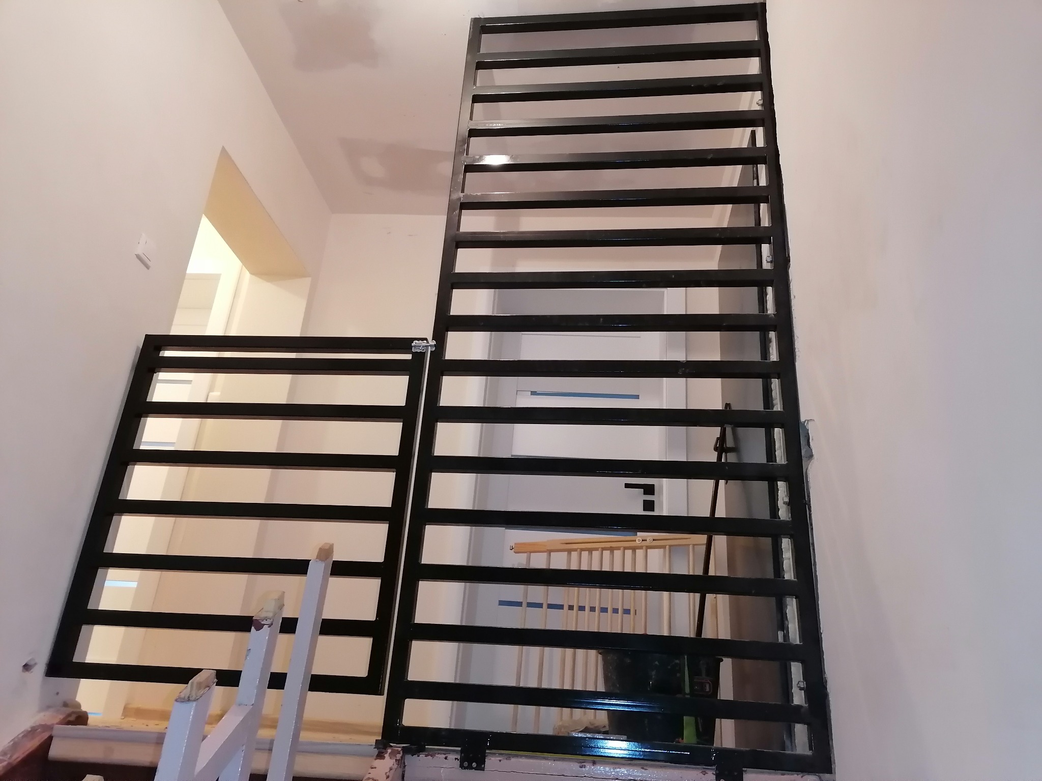 Częściowo zamontowana, czarna, metalowa konstrukcja schodowa z poziomymi szczeblami, widoczna na tle białej ściany i schodów w trakcie remontu.