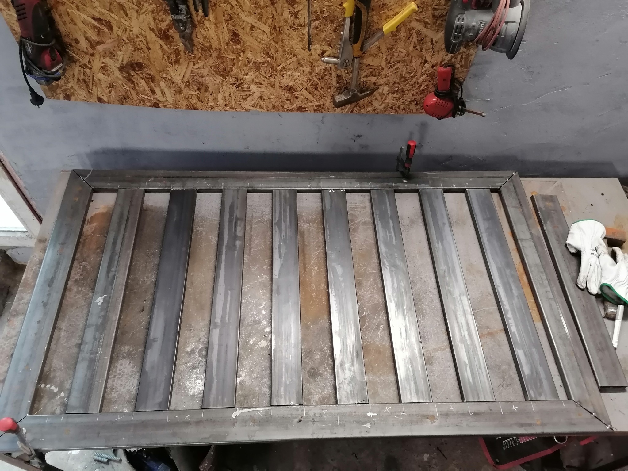 Metalowa konstrukcja z prostokątnych profili w trakcie spawania, widoczne zaznaczenia do spawania, w tle narzędzia i rękawice robocze.