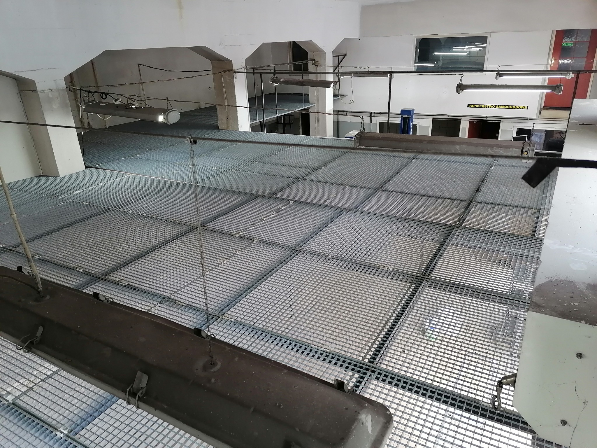 Wnętrze hali z metalową podłogą kratową, widok z góry na konstrukcję z oświetleniem zawieszonym na łańcuchach. W tle widoczne pomieszczenia na różnych poziomach oraz okno z napisem.