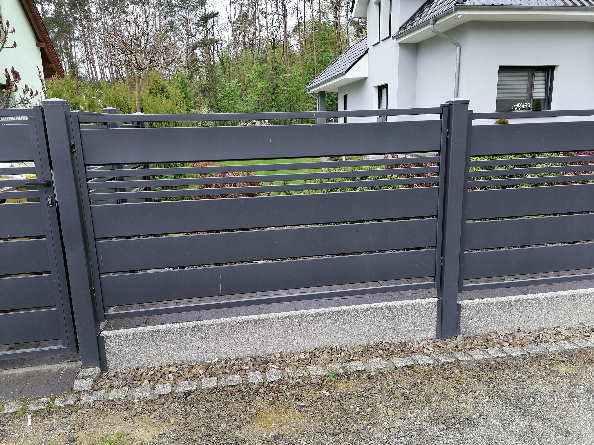 Nowoczesne, poziome ogrodzenie panelowe w kolorze grafitowym, z widocznym fragmentem domu w tle i elementami dekoracyjnymi z kory drzewnej u podstawy.