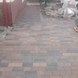 Ułożona kostka brukowa w odcieniach brązu i szarości, widoczna zagęszczarka w tle, częściowo zakończone prace przy budynku.