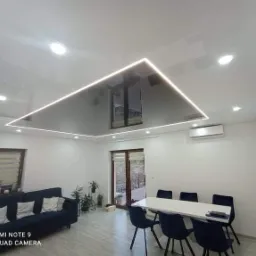 Nowoczesny salon z błyszczącym sufitem podwieszanym z oświetleniem LED w kształcie prostokąta, widoczny narożnik, stół z krzesłami oraz klimatyzator.