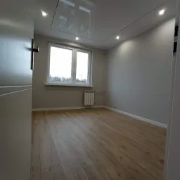 Pusty pok&oacute;j po remoncie z ułożonymi panelami podłogowymi w odcieniach brązu, szare ściany, białe okno z grzejnikiem i podwieszany sufit z oświetleniem punktowym.