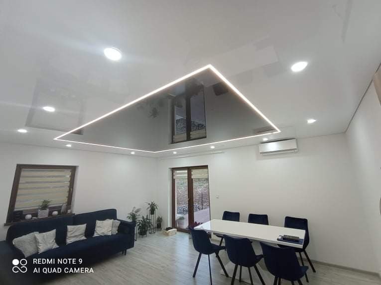 Nowoczesny salon z błyszczącym sufitem podwieszanym z oświetleniem LED w kształcie prostokąta, widoczny narożnik, stół z krzesłami oraz klimatyzator.