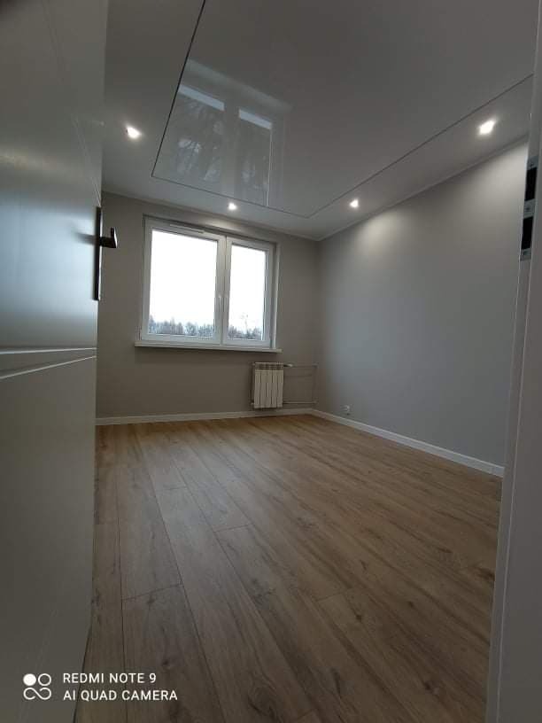 Pusty pokój po remoncie z ułożonymi panelami podłogowymi w odcieniach brązu, szare ściany, białe okno z grzejnikiem i podwieszany sufit z oświetleniem punktowym.