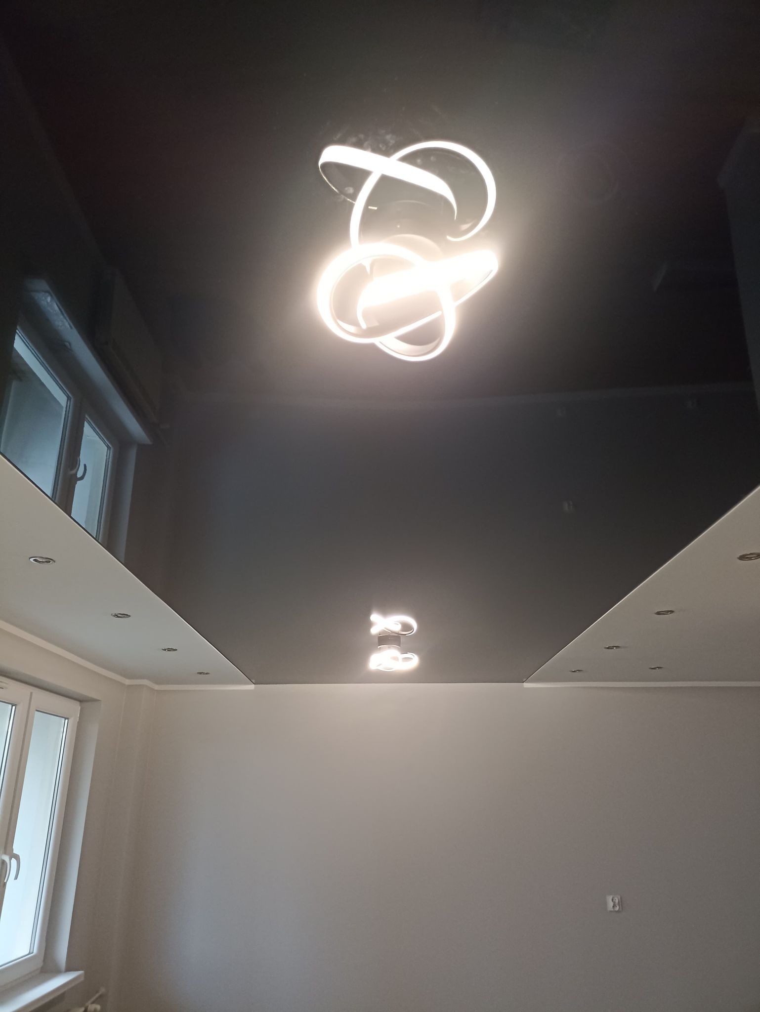 Nowoczesne oświetlenie LED w kształcie abstrakcyjnych okręgów, zamontowane na czarnym, lustrzanym suficie podwieszanym w jasnym pokoju z oknami.