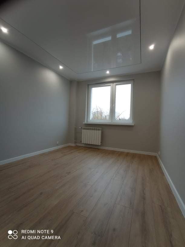 Wykończone wnętrze pokoju z panelami podłogowymi o wzorze drewna, szarą ścianą, białym sufitem podwieszanym z wbudowanym oświetleniem punktowym i grzejnikiem pod oknem.