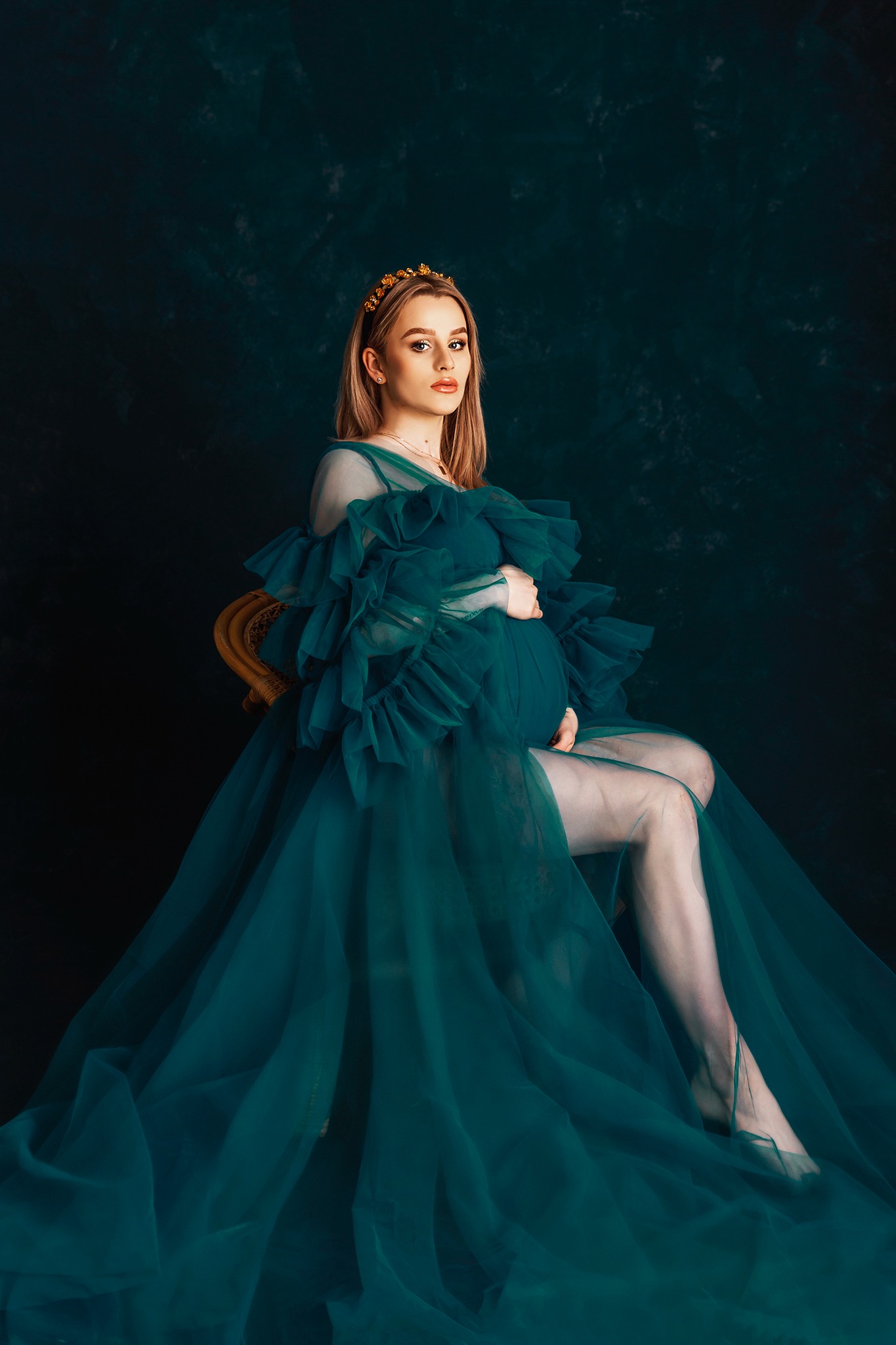 Portret kobiety w ciąży, siedzącej na krześle w zielonej, zwiewnej sukni z falbanami i złotej koronie na tle ciemnego, teksturowanego tła.