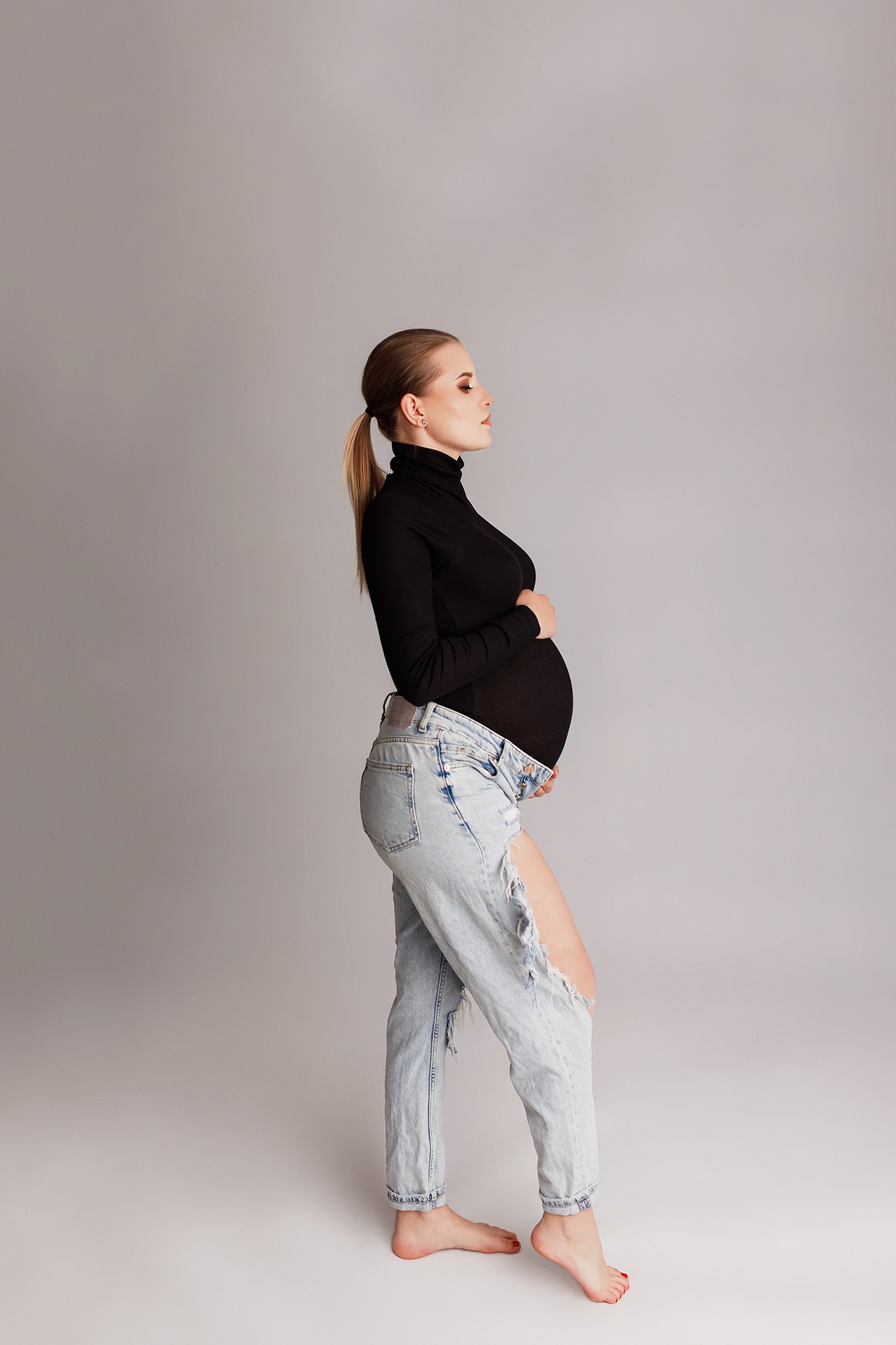 Kobieta w ciąży, w czarnym golfie i podartych jeansach, pozuje na szarym tle, widok z profilu, włosy spięte w kucyk, delikatny makijaż.