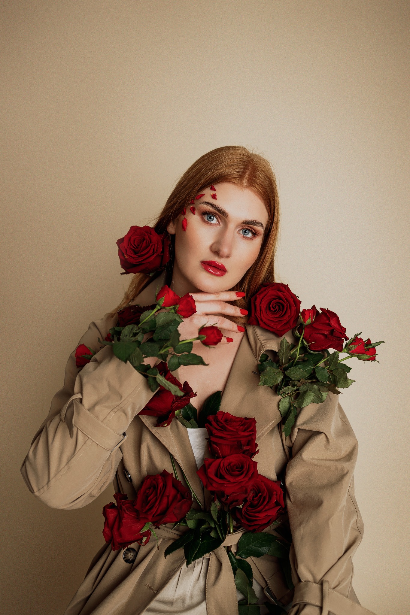 Portret kobiety o rudych włosach i niebieskich oczach w beżowym płaszczu, udekorowanej czerwonymi różami i płatkami róż na twarzy, z czerwonymi ustami i paznokciami.