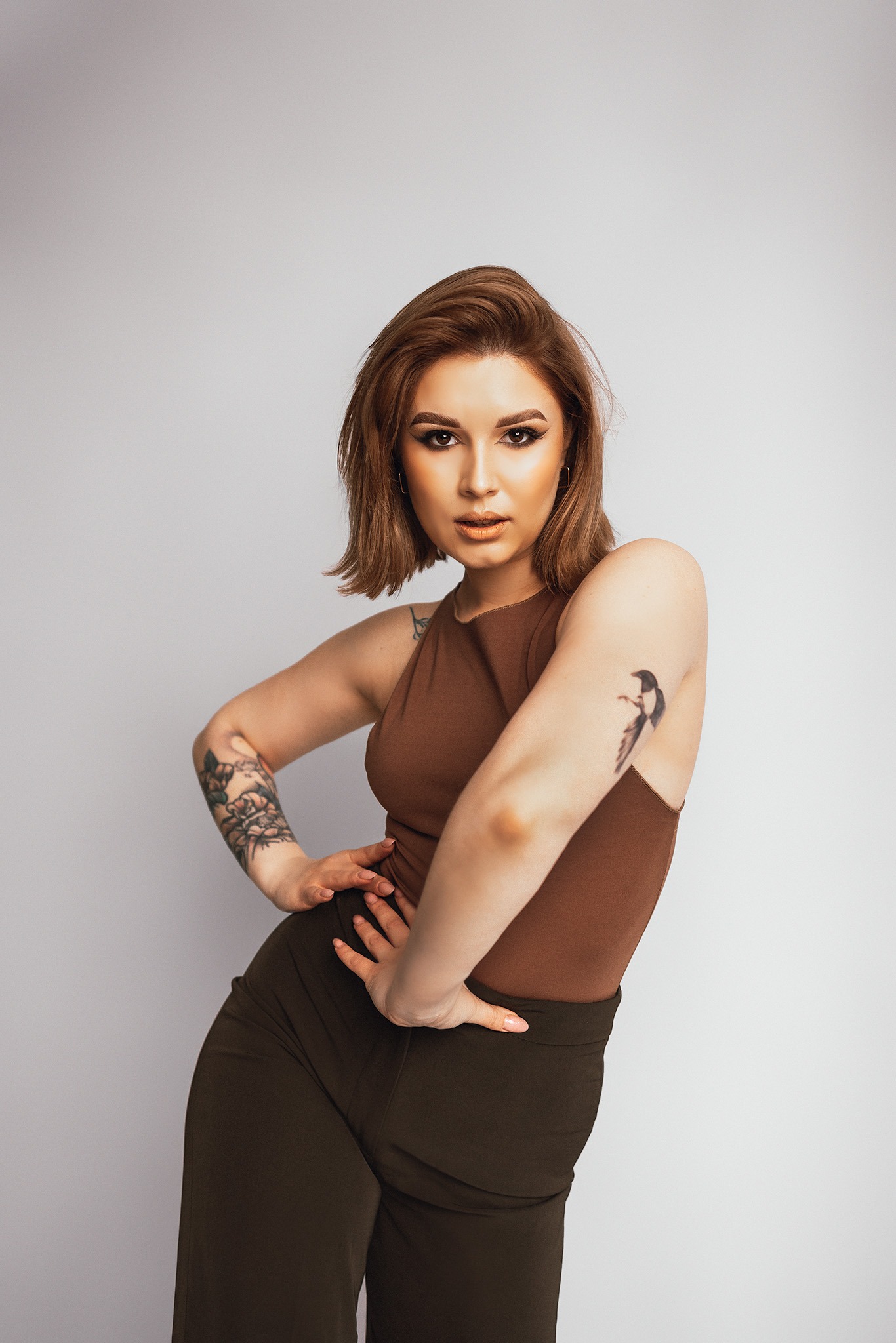 Portret kobiety z brązowymi włosami, w brązowym topie i ciemnozielonych spodniach, pozującej na jasnym tle z tatuażami na ramionach.