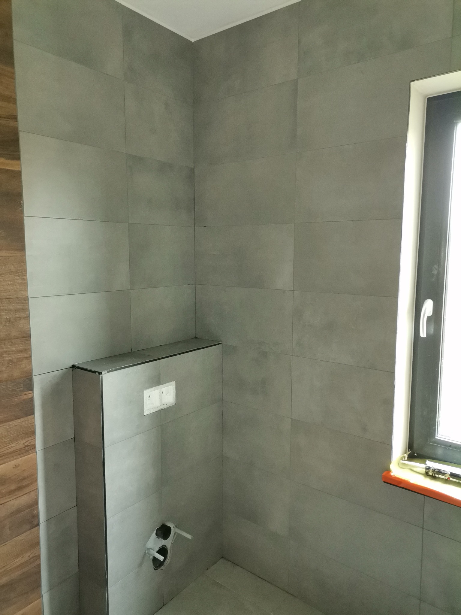 Szare, matowe płytki wielkoformatowe na ścianach łazienki, zabudowa stelaża WC, okno z czarną ramą, widoczne przyłącza wod-kan.