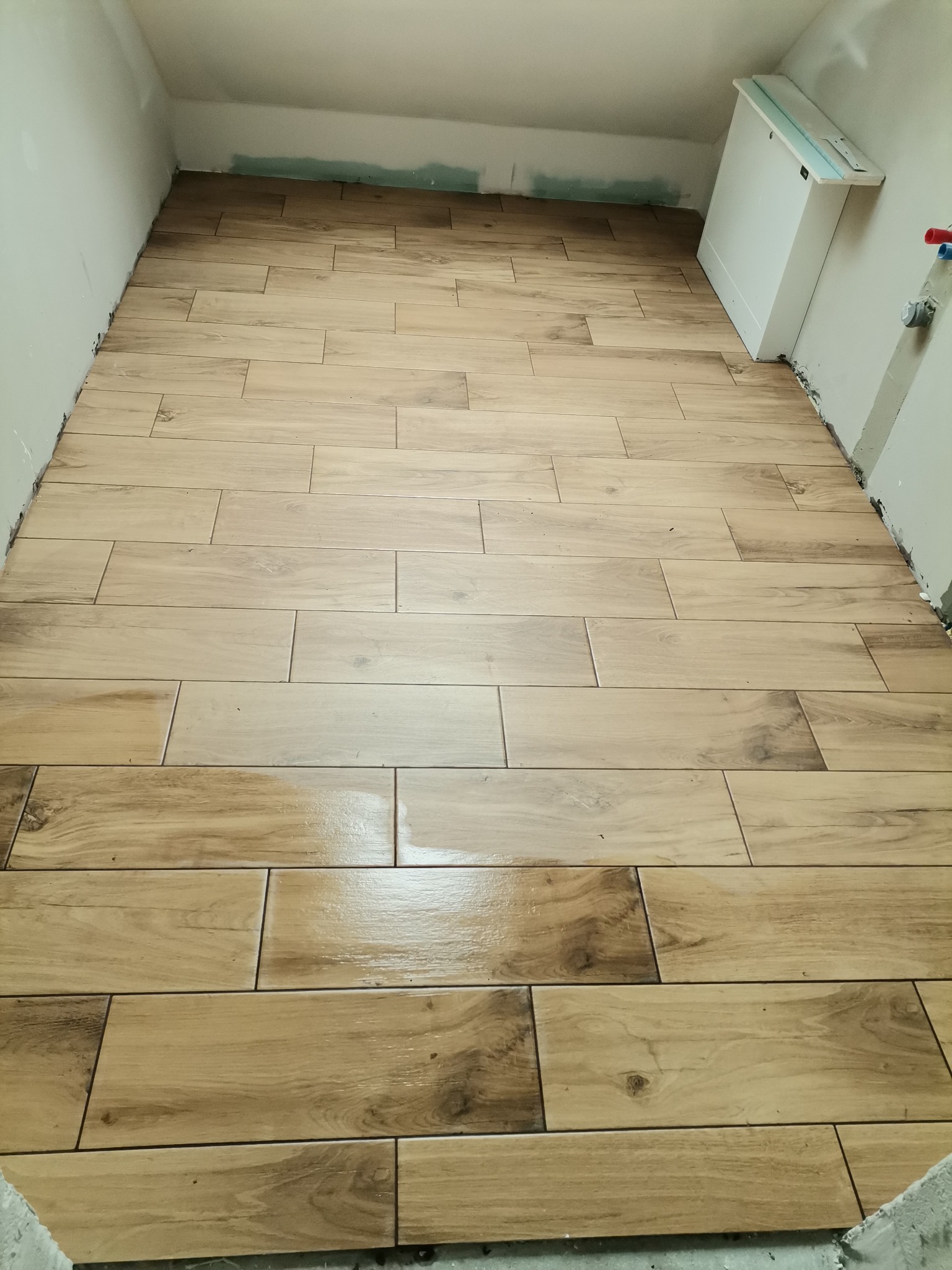 Ułożona podłoga z płytek imitujących drewno w łazience na poddaszu, widoczne niedokończone wykończenia ścian i zabudowa geberit.