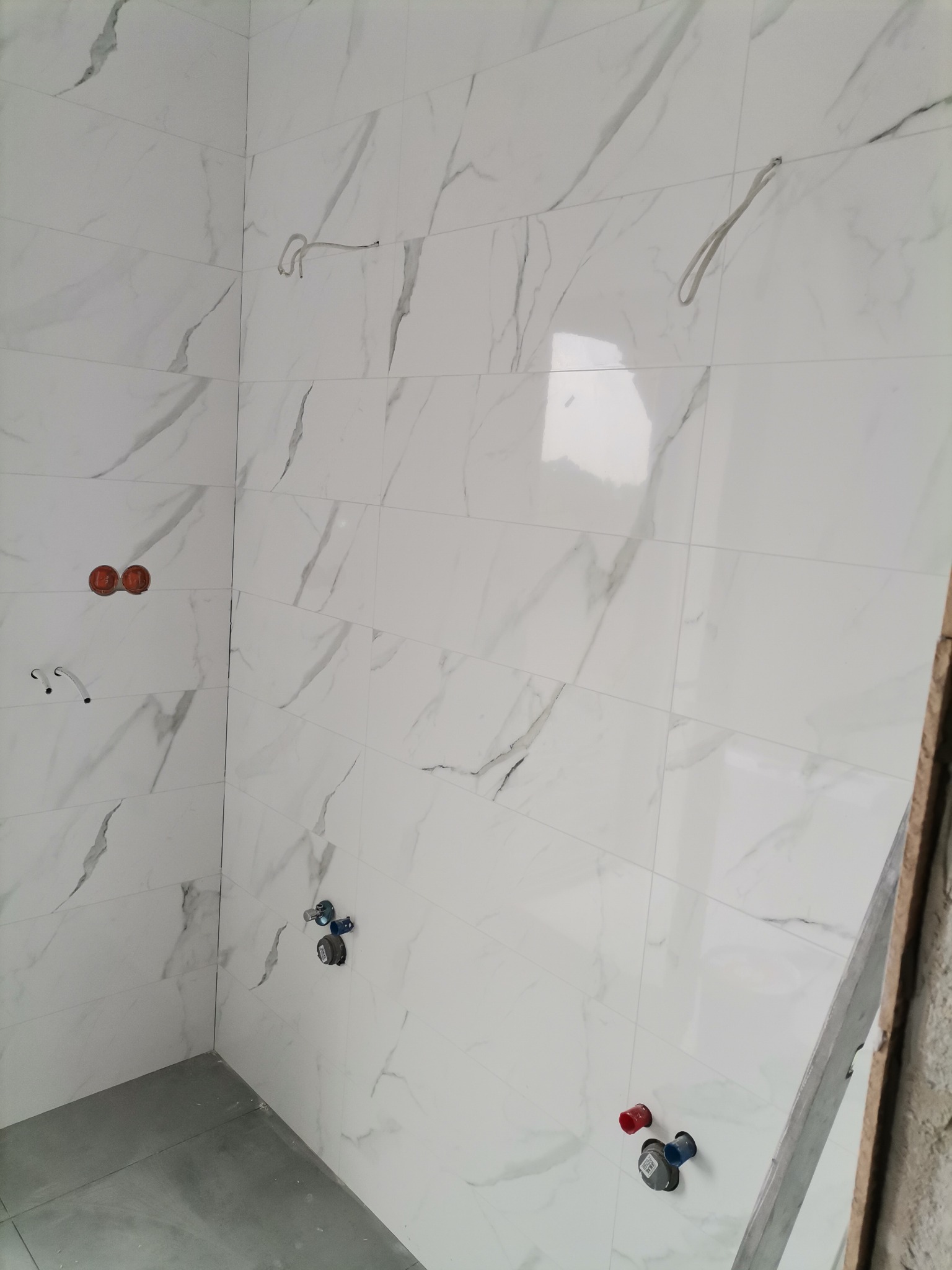 Ściany łazienki wyłożone białymi płytkami imitującymi marmur, widoczne wyprowadzenia instalacji wodnej i elektrycznej przed montażem armatury i oświetlenia, szare płytki podłogowe.