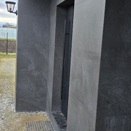 Doklejanki - Nowoczesna elewacja budynku z ciemnoszarych płytek, z widoczną lampą zewnętrzną, matą wejściową i systemem alarmowym.