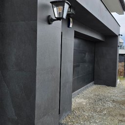 Doklejanki - Nowoczesna elewacja garażu wykończona grafitowymi płytami, z zamontowaną lampą zewnętrzną o klasycznym kształcie i czujnikiem ruchu