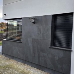 Doklejanki - Szara elewacja domu z oknami z roletami zewnętrznymi i lampą elewacyjną, widok z poziomu gruntu.