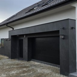 Doklejanki - Nowoczesny dom z czarną bramą garażową i elewacją z ciemnego kamienia, oświetlony zewnętrznymi lampami, widok na dach z dachówką.