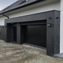 Nowoczesna elewacja domu z garażem, wykończona ciemnoszarymi płytami elewacyjnymi i czarną bramą garażową, oświetlona lampami zewnętrznymi.