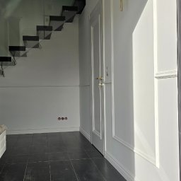 Doklejanki - Nowoczesne wnętrze: czarna podłoga z płytek, białe ściany z dekoracyjnymi listwami, schody z szklaną balustradą i designerski kinkiet.