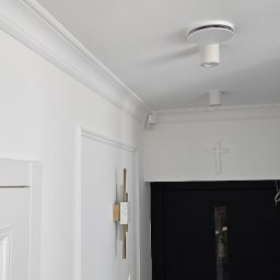 Doklejanki - Białe wnętrze z dwoma okrągłymi lampami sufitowymi, listwami dekoracyjnymi pod sufitem, kinkietem z elementami złota i marmuru oraz krzyżem nad czarnymi drzwiami.