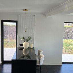 Doklejanki - Minimalistyczna kuchnia z czarnym blatem wyspowym, białymi ścianami z dekoracyjnymi panelami pionowymi, czarną podłogą i jasnymi krzesłami barowymi.