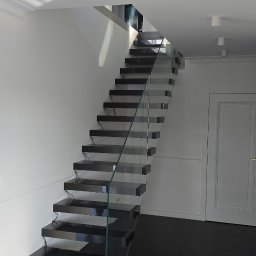 Doklejanki - Nowoczesne schody z czarnego kamienia z przezroczystą szklaną balustradą w jasnym wnętrzu z białymi ścianami i ciemną podłogą.