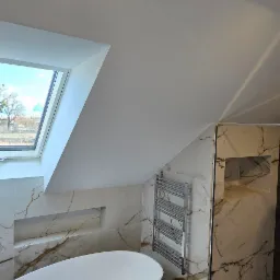 Nowoczesna łazienka na poddaszu z owalną wanną, grzejnikiem drabinkowym i kabiną prysznicową wykończoną marmurowymi płytkami z żyłkowaniem, doświetlona oknem dachowym.