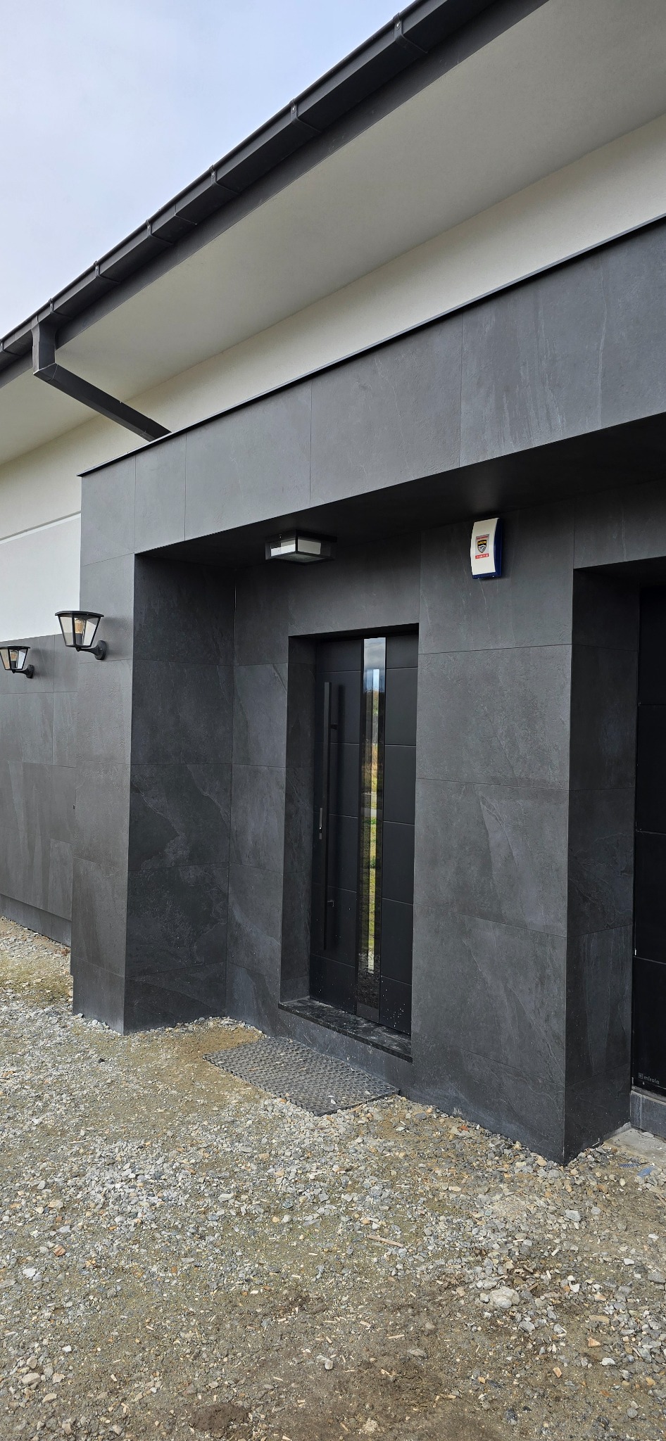 Nowoczesne wejście do domu z elewacją z ciemnoszarych płyt imitujących kamień, czarne drzwi z pionowymi przeszkleniami, oświetlenie zewnętrzne oraz system alarmowy.