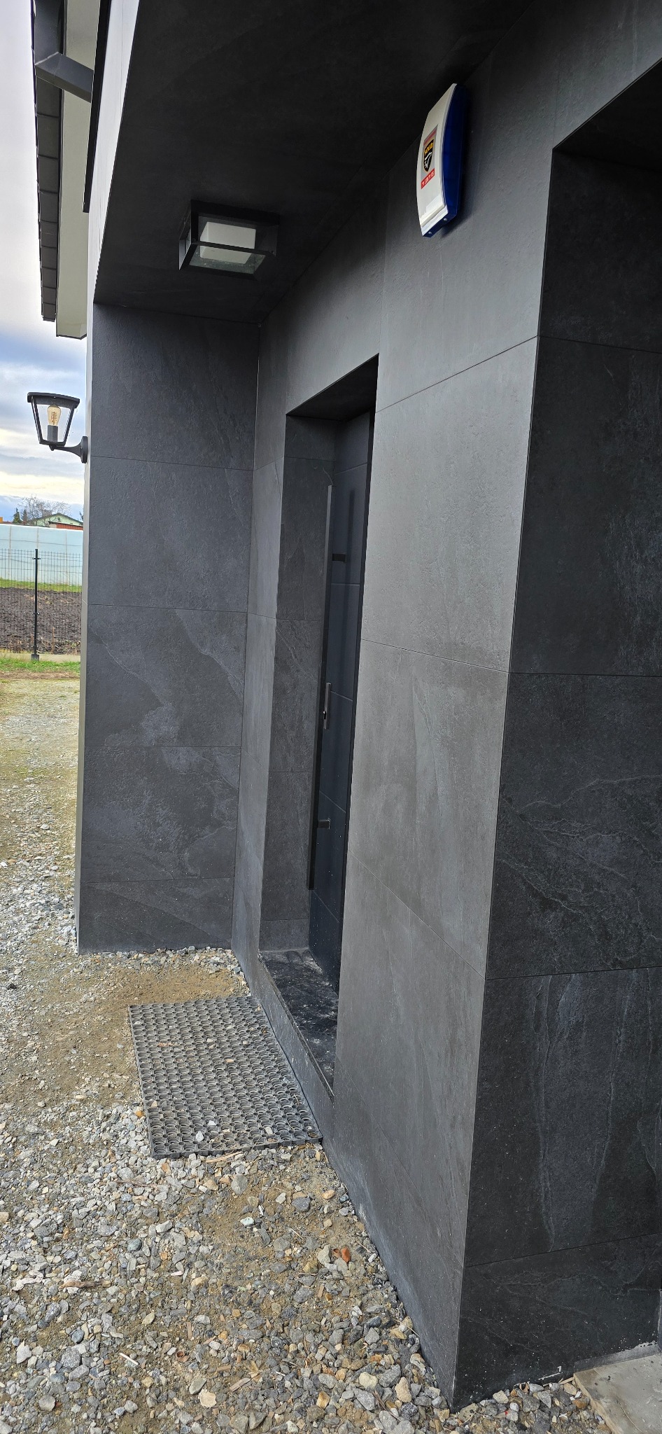 Nowoczesna elewacja budynku z ciemnoszarych płytek, z widoczną lampą zewnętrzną, matą wejściową i systemem alarmowym.