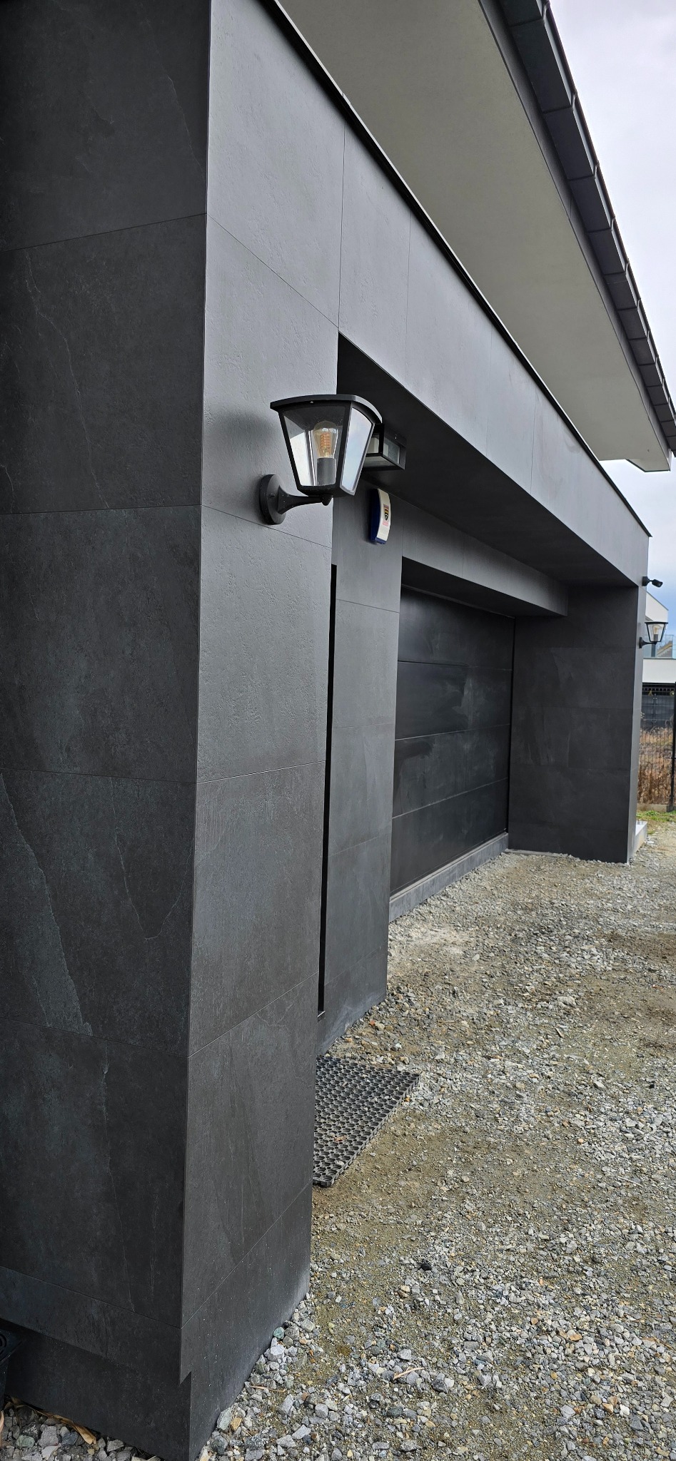 Nowoczesna elewacja garażu wykończona grafitowymi płytami, z zamontowaną lampą zewnętrzną o klasycznym kształcie i czujnikiem ruchu