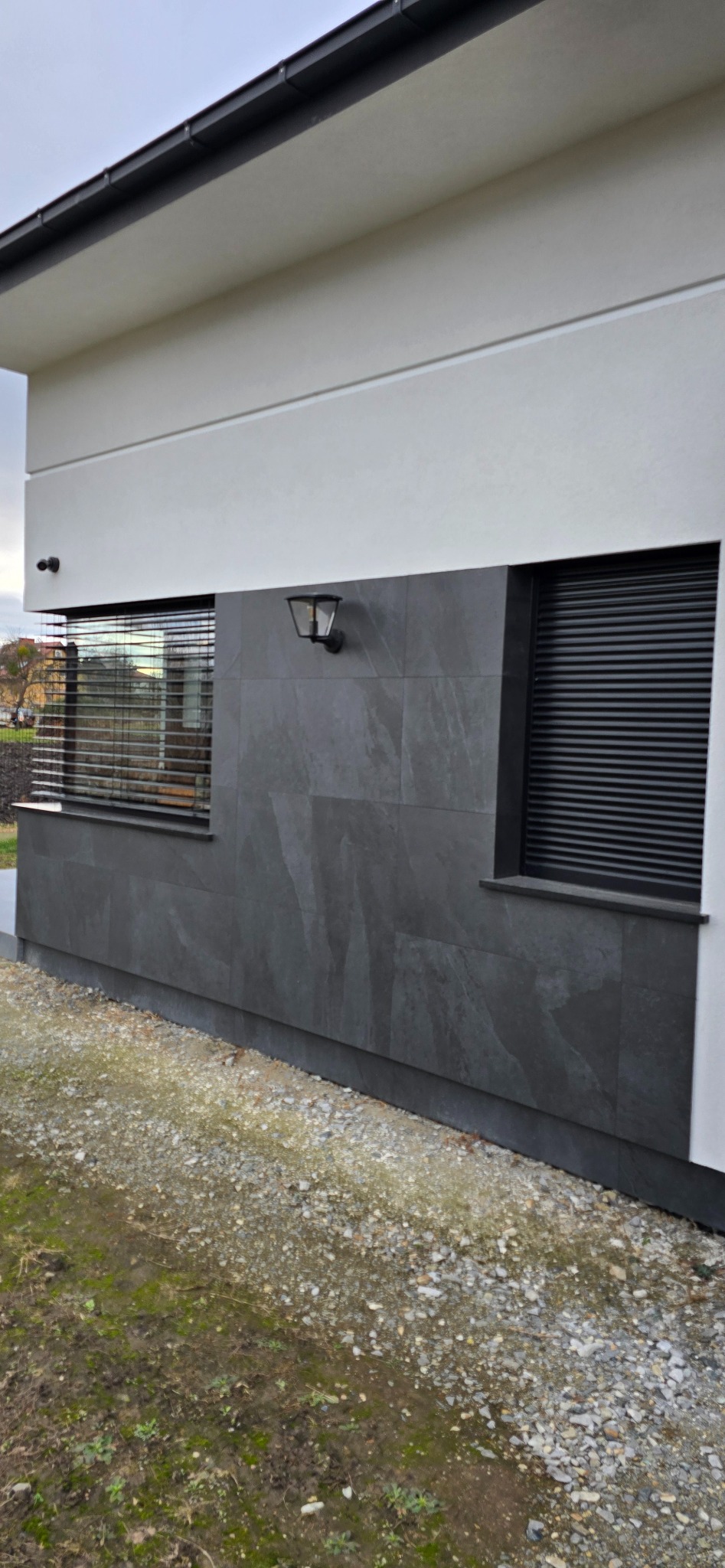 Szara elewacja domu z oknami z roletami zewnętrznymi i lampą elewacyjną, widok z poziomu gruntu.