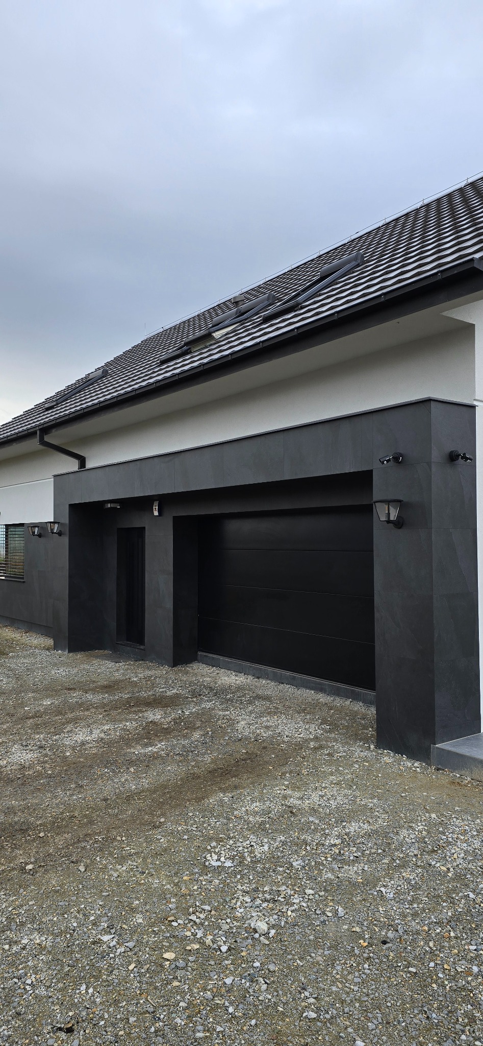 Nowoczesny dom z czarną bramą garażową i elewacją z ciemnego kamienia, oświetlony zewnętrznymi lampami, widok na dach z dachówką.