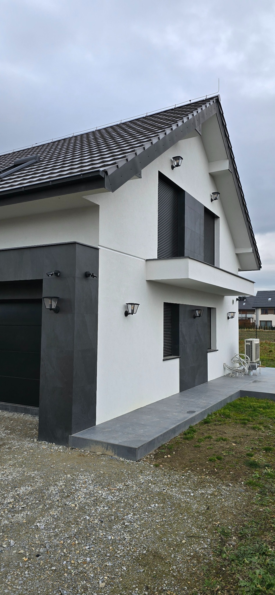 Nowoczesny dom jednorodzinny z elewacją w kolorach bieli i grafitu, widoczne okna z żaluzjami zewnętrznymi, oświetlenie elewacyjne oraz wykończony chodnik z szarej kostki.