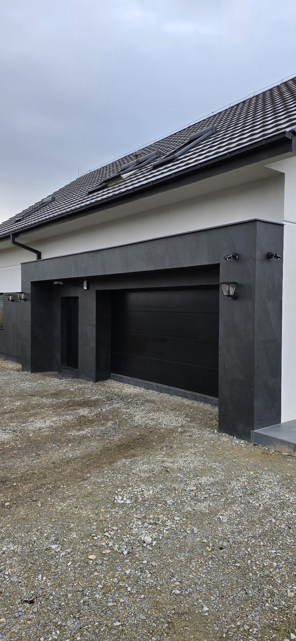 Nowoczesna elewacja domu z garażem, wykończona ciemnoszarymi płytami elewacyjnymi i czarną bramą garażową, oświetlona lampami zewnętrznymi.