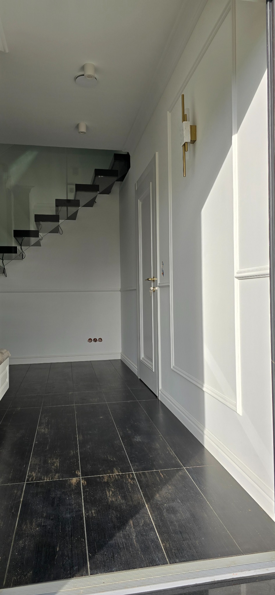 Nowoczesne wnętrze: czarna podłoga z płytek, białe ściany z dekoracyjnymi listwami, schody z szklaną balustradą i designerski kinkiet.