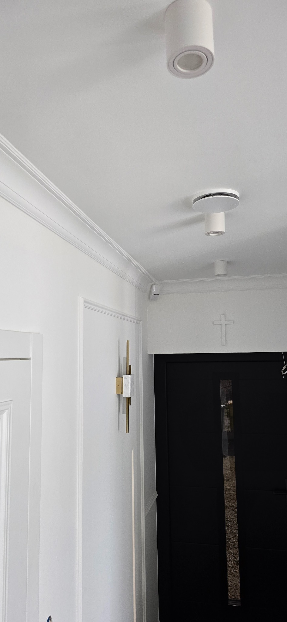 Białe wnętrze z dwoma okrągłymi lampami sufitowymi, listwami dekoracyjnymi pod sufitem, kinkietem z elementami złota i marmuru oraz krzyżem nad czarnymi drzwiami.