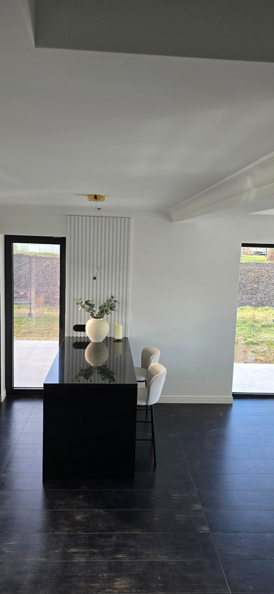 Minimalistyczna kuchnia z czarnym blatem wyspowym, białymi ścianami z dekoracyjnymi panelami pionowymi, czarną podłogą i jasnymi krzesłami barowymi.